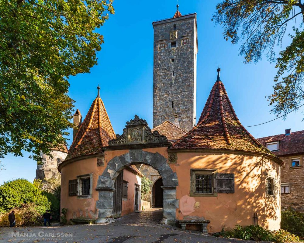 Rothenburg ob der Tauber - Zusehen ist das dickbäuchige Burgtor in organge mit dem hohen kantigen Burgturm und Stadttor aus alten grauen Steinen. Das Sonnenlicht scheint quer von rechts auf den Turm. Vom vorderen Turm sieht man den Durchgang und die Brücke zum hinteren Turm.